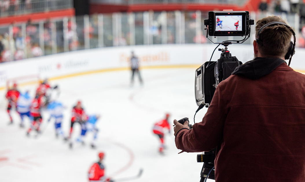 A cameraman captures the action of an ice hockey game, focusing on the rink with players in red and blue jerseys, while the match is displayed on the camera screen.